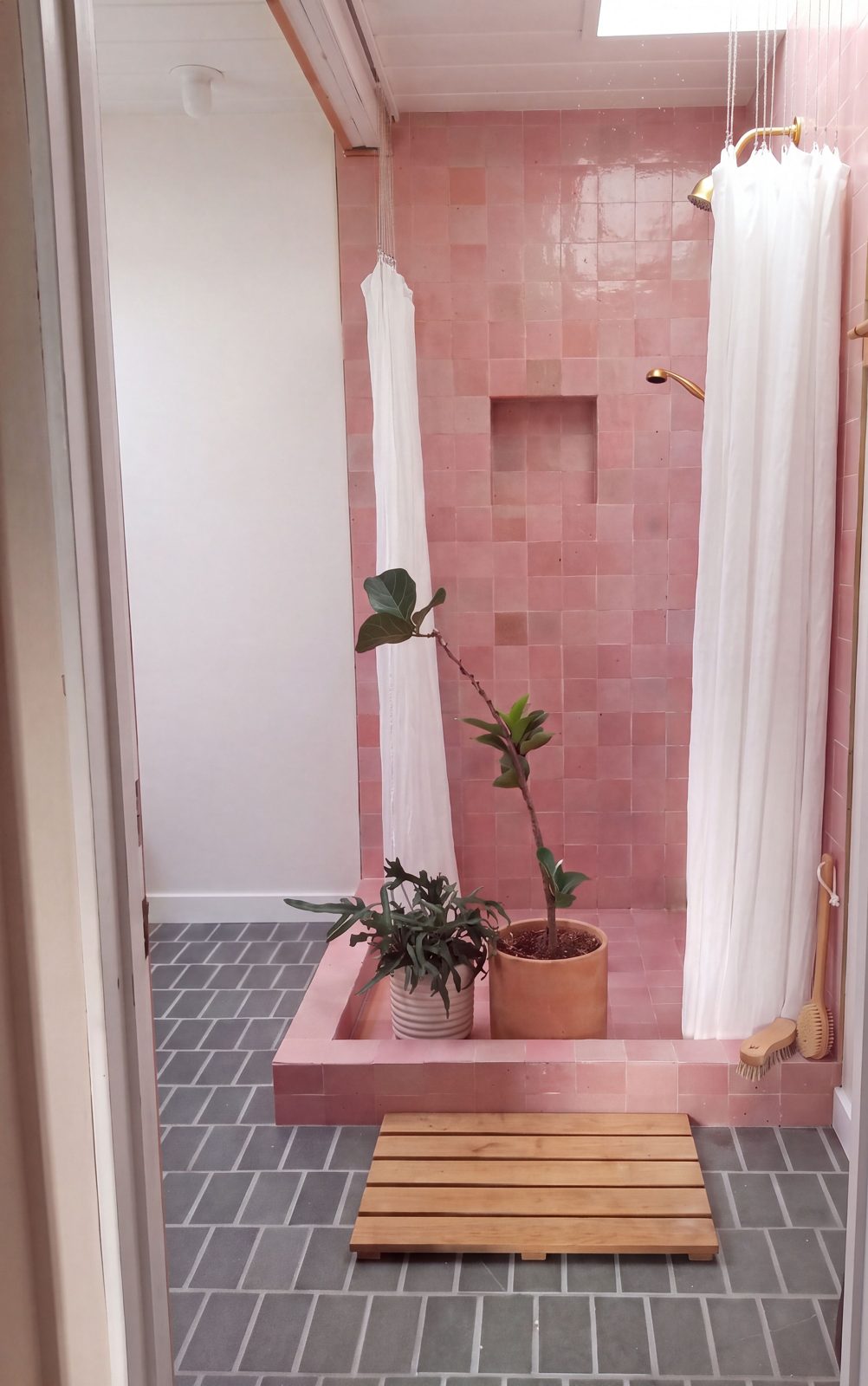 Bright bathroom with a pink tile shower, plants and a wood bath mat